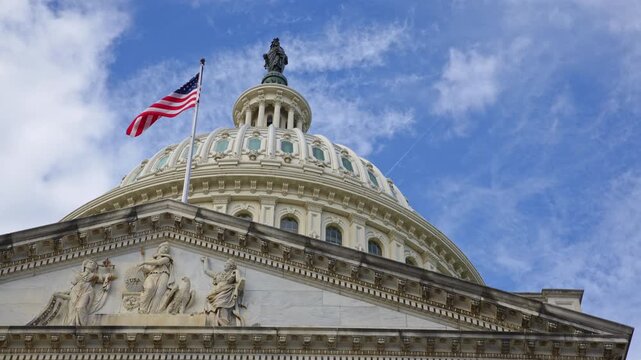 American flag waving strongly on Capitol. American flag above clouds over US Capitol. American flag enduring symbol of Amerika freedom. American flag on historic Capitol dome in Washington DC.