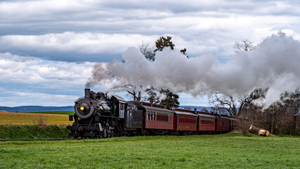 A steam train moves through green fields with trees on the side. Dark smoke rises from the engine as it pulls red passenger cars along the tracks on a cloudy day.