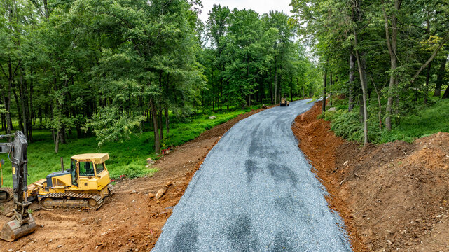 Heavy machinery is working on a new road that is being built through a forested area. Workers are moving earth and laying gravel on the ground. Trees surround the site.