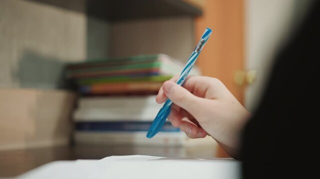 Hand grasps pen. Individual holding pen while studying in peaceful environment. Serene setting featuring hand gripping blue pen over notebook amidst warm lighting and bookshelf background