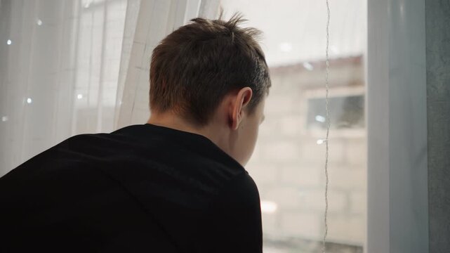 Child looks out. Boy observes outside from behind window. Child gazes through rain speckled glass at outdoors. Young silhouette of boy peering through rainspeckled window into urban backyard