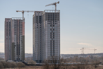 Fototapeta premium Three cranes beside rising residential blocks, skeletal floors, window openings, pale sky, remote operator cabin, urban planner reviewing blueprint at roadside, muted winter light