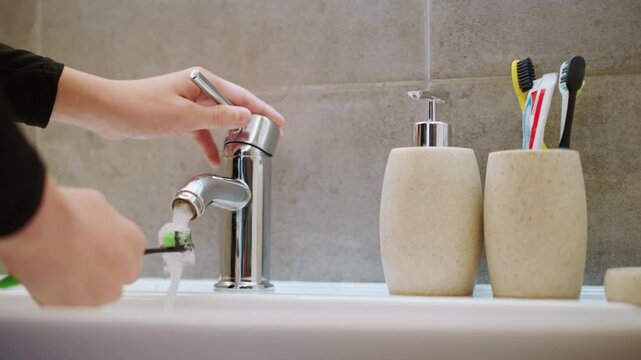 Grandma helping grandson brush teeth, closeup of hands turning chrome faucet, toothbrushes in stone holders, water rinsing bristles, soft morning light, warm home atmosphere, gentle caregiving