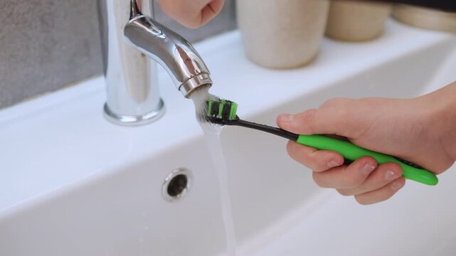 Green toothbrush under chrome faucet rinsing, hand holding toothbrush under running water over porcelain sink, soap dispensers on counter, bristles wet and dripping, morning family hygiene, gentle