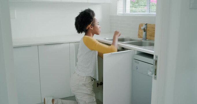 Woman kneeling opening cabinet under sink in kitchen, inspecting plumbing and using toolkit