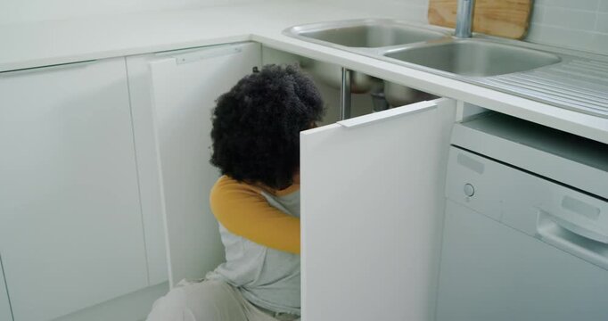 African American woman opening cabinet, checking, fixing pipes under kitchen sink using tools