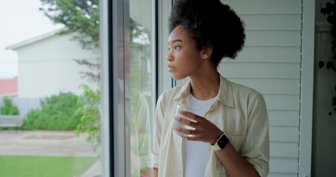 African woman standing by glass door lifting glass, sipping, scanning yard, contemplating view