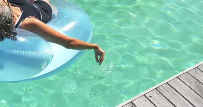Adult African woman in swimsuit reclining on ring extending arm skimming water for rest near dock