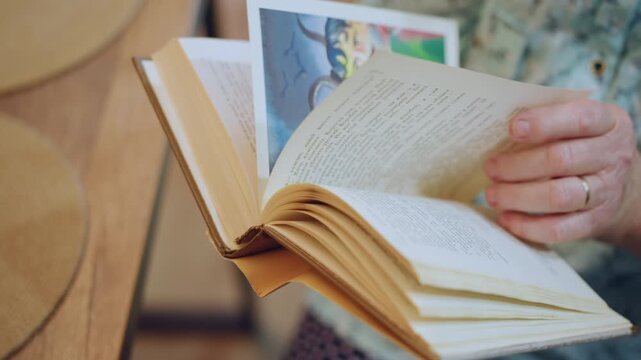 Open book with illustration pages, grandma gently revealing colorful picture while reading aloud, hands showing steady thumb and ring, storytime vibe in sunlit kitchen, turning pages to childlike art,