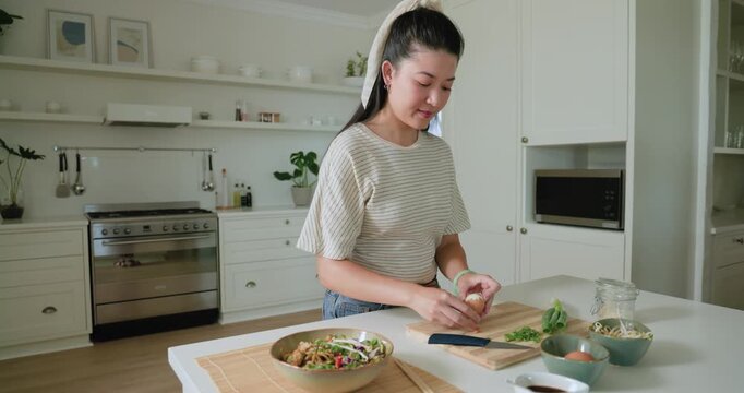 Adult Asian woman reaching for egg, peeling shell and preparing salad on board at kitchen island