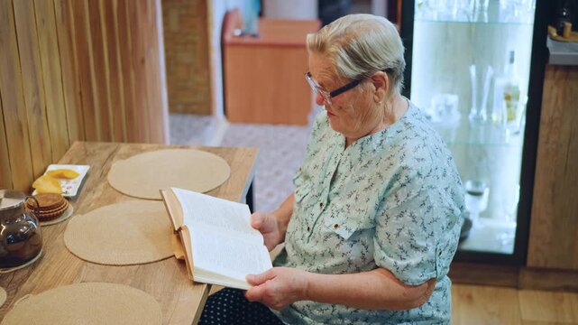 Grandma reads peacefully. Elderly grandmother immersed in book amidst breakfast setting with floral attire. An older woman leisurely reads her book at breakfast table surrounded by homey details