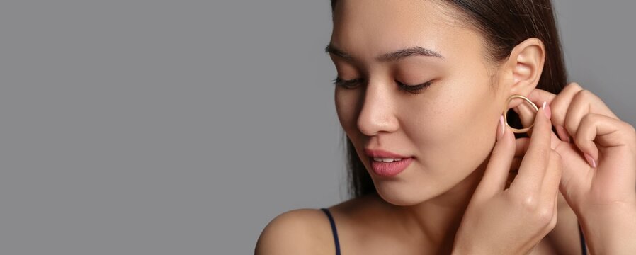 Young Asian woman putting on beautiful earrings against grey background with space for text