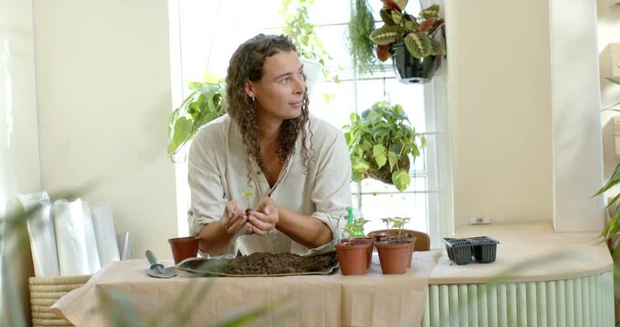 African nonbinary adult preparing seedling for potting, moving soil with trowel at table by window