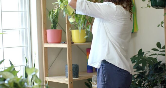 Nonbinary adult tending hanging plant on shelf by window, inspecting then misting with sprayer