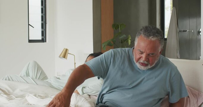 Alarm clock sounding African American couple in pajamas waking and making bed with duvet in bedroom