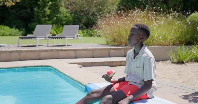 Child boy cooling down in sunny backyard pool on mat, dipping feet, eating watermelon, copy space