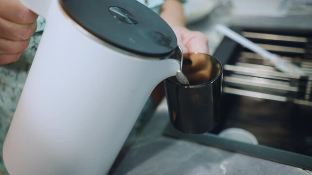 White grandmother pouring tea into glass mug from white kettle, careful steady pour with steam swirl, dark green cup on marble countertop, quiet cozy finish to morning ritual, attentive homemaker