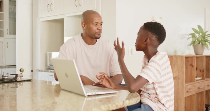 Father beige tee, teen son striped tee gesturing at laptop, father explaining homework in kitchen