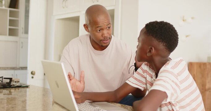 Explaining father in beige tee guiding son in striped tee at kitchen island, using laptop keyboard