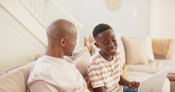 African father and teen son sitting on couch, son turning from laptop, explaining, father listening
