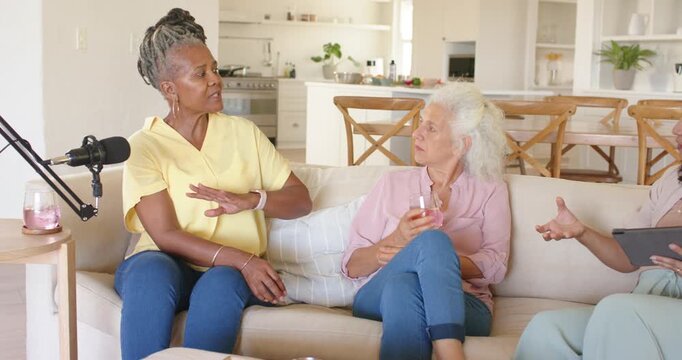 Diverse female cohosts on sofa checking tablet notes after left host leaning and gesturing, mic