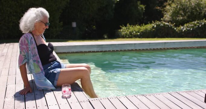 Shifting senior woman dipping feet to cool from sun, kimono on yard pool, glass, copy space