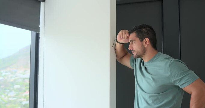 Adult male leaning on white partition at home window, gazing out then rubbing forehead, copy space