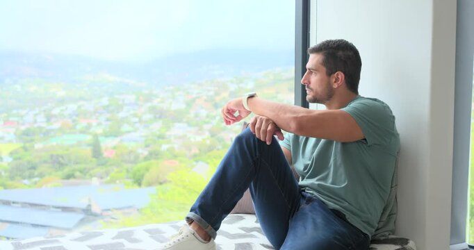 Man 20s-40s sitting on window seat, watching hills through large window, rubbing nose, copy space