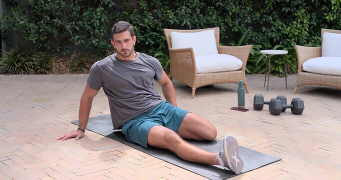 Grey-shirted man shifting, hugging right knee for hip stretch on patio mat beside dumbbells, bottle