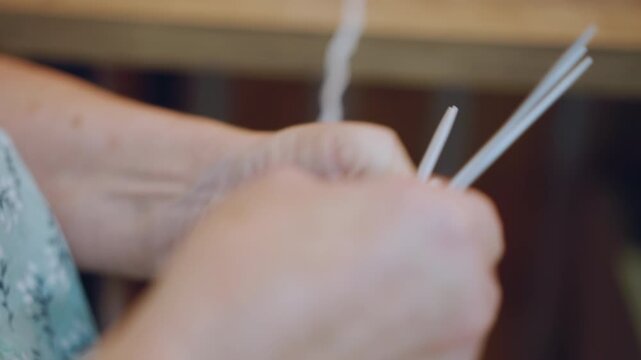 Closeup elderly white grandma knitting hands, slow patient fingers looping yarn around metal needles, soft wool texture, wrinkled hands showing decades of practice, calm domestic atmosphere in cozy