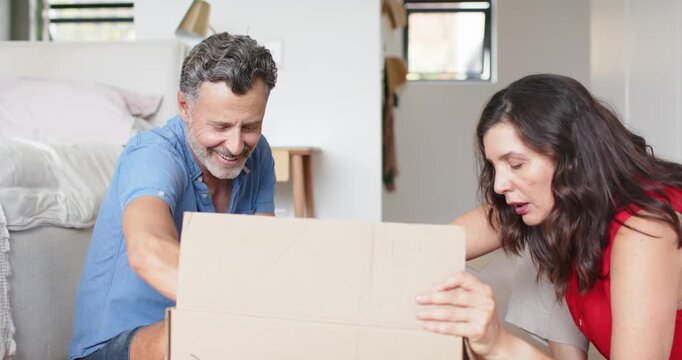Mature couple unpacking moving box while man lifting pink sweater and woman placing hand to chest