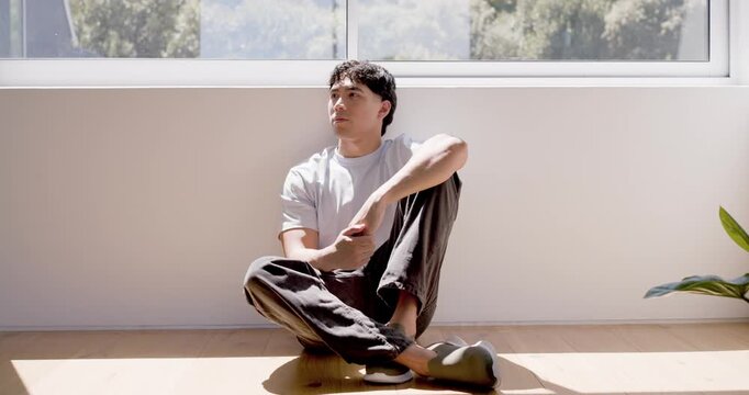 Asian man sitting round cushion under wide window, sun patch drawing gaze, turning to face camera