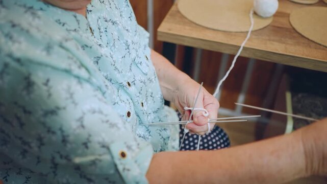 Elderly caucasian woman knitting near table, medium framing shows ball of yarn, multiple needles and looping hands creating slow rhythmic stitches, cozy domestic setting and sense of generational
