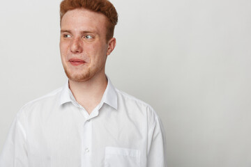 Obraz premium Young man with red hair and freckles wearing white shirt looks sideways with thoughtful expression on plain light background. Concept of casual style, contemplation, and natural appearance.