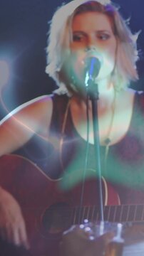 Vertical video: Hand with glass revealing musician strumming guitar, performing at mic under flares