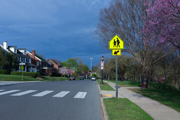 Residential neighborhood in Frederick, Maryland, with yellow school crossing sign and pink spring blossoms under a dark storm sky on S Jefferson Street. © Alexey Konkov
