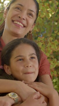 Vertical video: Hugging mother sharing love with girl under tree, sunlight flaring, watch, rust top