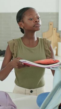 Vertical video: Packing sister in olive top fitting books and pouch into bag at counter for school