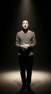 Sad mime stands under a spotlight on a dark stage.