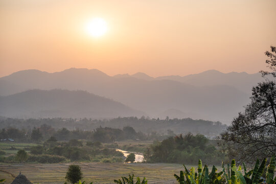 Exotic rural plantations and rice fields landscape of Pai city and river in Northern Thailand with fog from forest fires covering hills and meadows at sunset, sun in smog shining at countryside.
