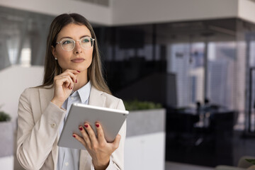 Thoughtful professional woman stands in modern office, holding digital tablet, staring away with pensive expression, looks in deep concentration or decision-making. Innovations and strategy planning © fizkes