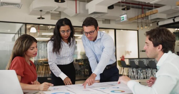Diverse agency staff analyzing paper marketing reports on meeting table, reviewing charts, brainstorming on project strategy, discussing research results, KPI benchmarks