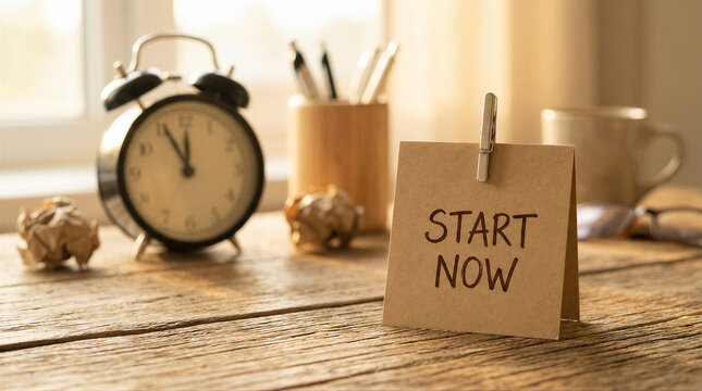 Motivational start now note on a rustic wooden desk with an alarm clock, crumpled paper, and office supplies, bathed in warm light