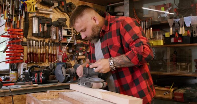 Carpenter occupation. Joinery process. Man smoothing board surface with belt sander in workshop interior with tools kit on shelves.