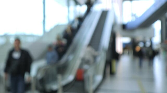Blurred airport scene. People ride escalators. Travelers walk around. Background lights glow. No animals or plants visible