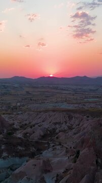 Vertical video. Goreme, Nevsehir, Turkey. Drone flight over Red Valley during sunset with sun disappearing behind mountains and lighting up rock formations.. Aerial View, Departure of the camera. Ric