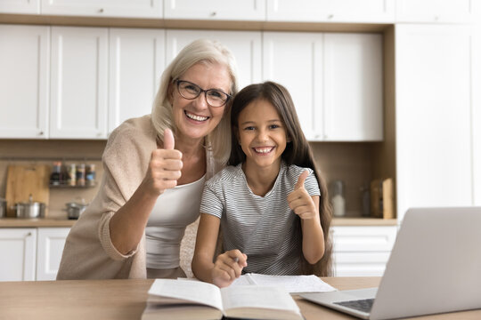 Portrait of girl and her older granny show thumb up. Cute little child studying online using laptop with help of grandmother in kitchen, smile, looking at camera. Effective learning, results, tuition