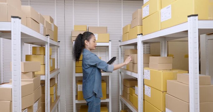 Asian woman entrepreneur slowly moves along storage aisle, points at parcel boxes while reviewing order list on tablet, manages warehouse inventory and online store fulfillment process