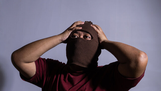 An adult male wearing a brown knit balaclava grabs his head with both hands in a dramatic pose of extreme frustration and stress. He looks upward with a sweaty face and tired eyes.