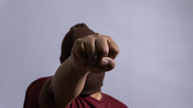 An adult male wearing a brown knit balaclava extends a clenched fist directly outward in a powerful and aggressive forward punch. The tight fist is perfectly sharp while the masked